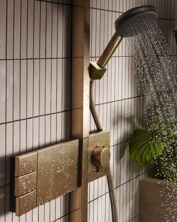 Hansgrohe handdouche in een warme metaaltint met stromend water, verticale wandtegels en een groen blad.