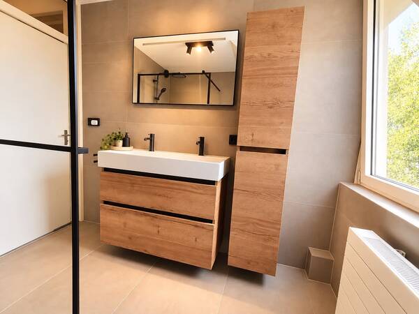 Badkamer met beige tegels, zwevend houtlook wastafelmeubel, hoge kast, zwarte kranen en een zwart omlijste spiegel.