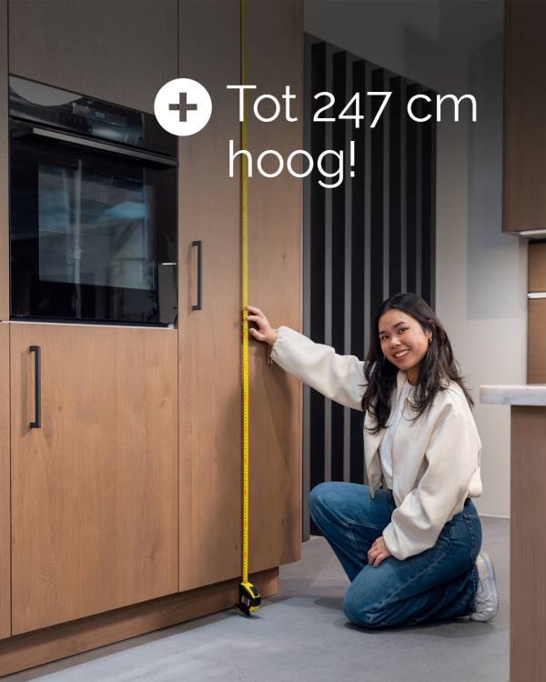 Vrouw meet een hoge keukenkast met houtlook, naast een zwarte inbouwoven. Tekst: tot 247 cm hoog.