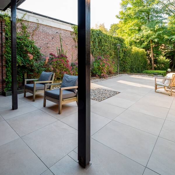 Terras met grote lichtgrijze tegels, houten stoelen, zwarte overkapping en groene beplanting langs een bakstenen muur.
