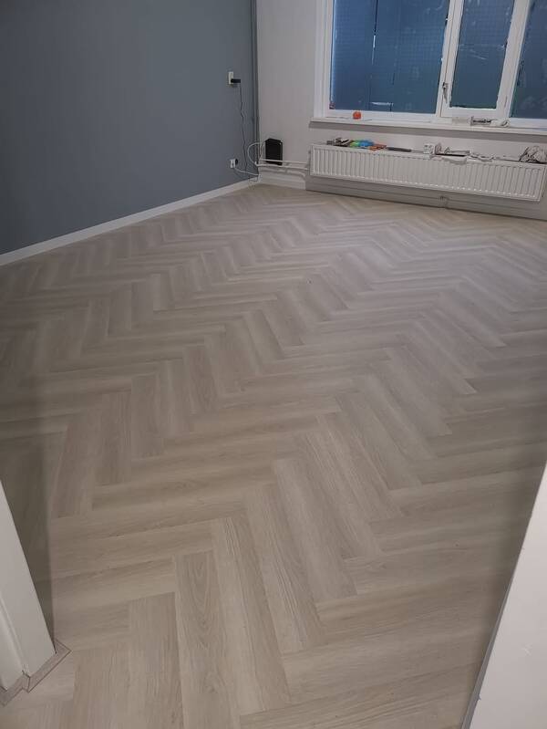 Lichte PVC-clickvisgraatvloer met houtlook in een ruimte met een blauwgrijze wand en witte plinten.
