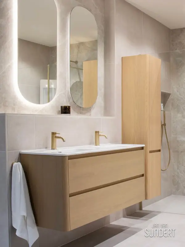 Badkamer met licht houten wastafelmeubel, zandkleurige tegels, afgeronde spiegels en champagnekleurige kranen.