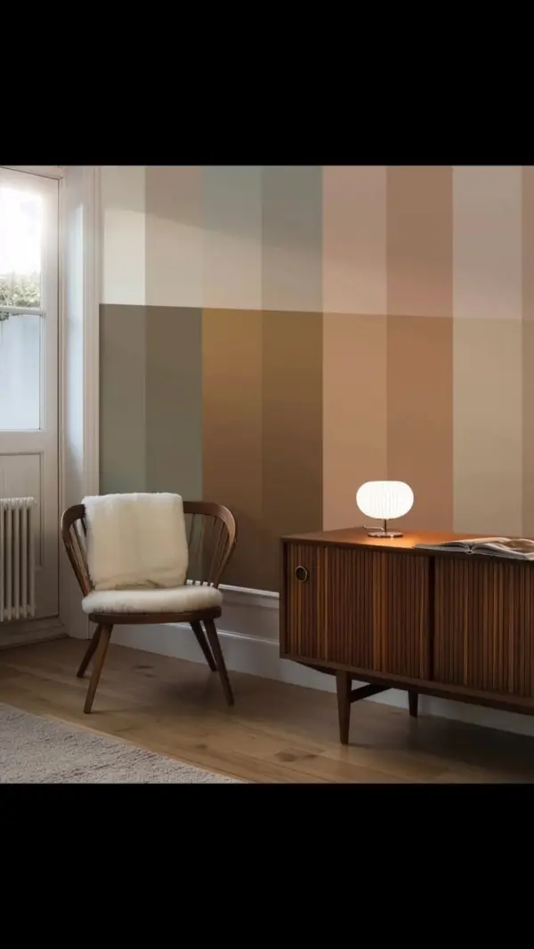 Behang met brede kleurvlakken achter een houten stoel met lichte bekleding en een donker dressoir met witte tafellamp.