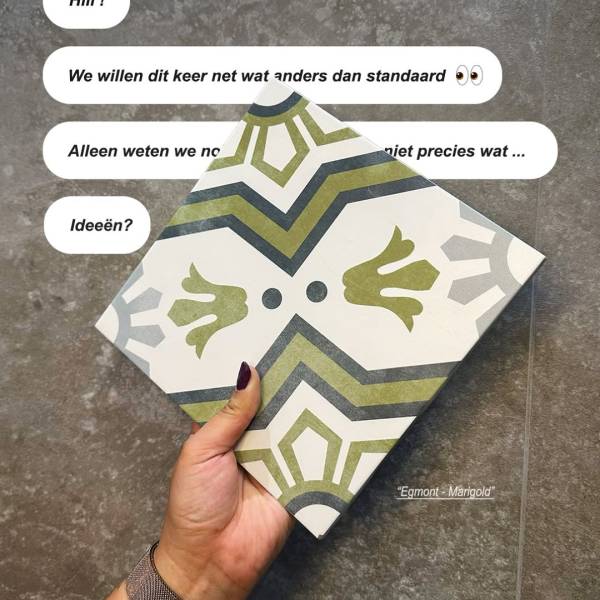 Een hand houdt de Egmont Marigold-tegel vast, met een groen en grijs decoratief patroon op een lichte basis.